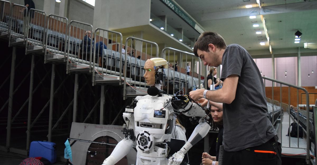 A technician works on a full-size humanoid robot on a stand at an indoor technology exhibition.
