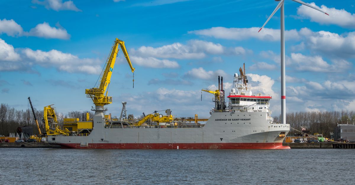 A pipe-burying vessel moored at a harbour quay with a wind turbine behind it